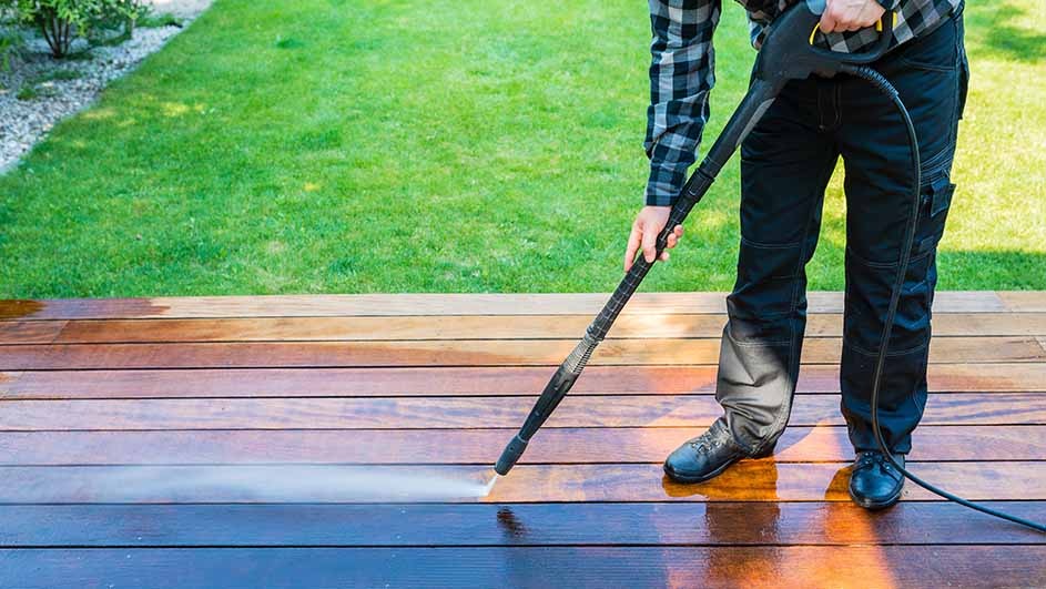 A man is cleaning a wooden terrace with a Kärcher pressure washer, making the surface look bright and fresh in a green garden.