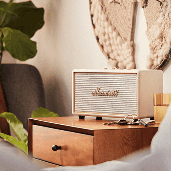 Een Marshall speaker staat op een houten nachtkastje, naast een glas en sleutels, met een plant en decoratie op de achtergrond.