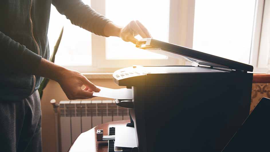 Een persoon vult papier in een zwarte printer met een open scannerbedekking, voor een raam en een verwarming.