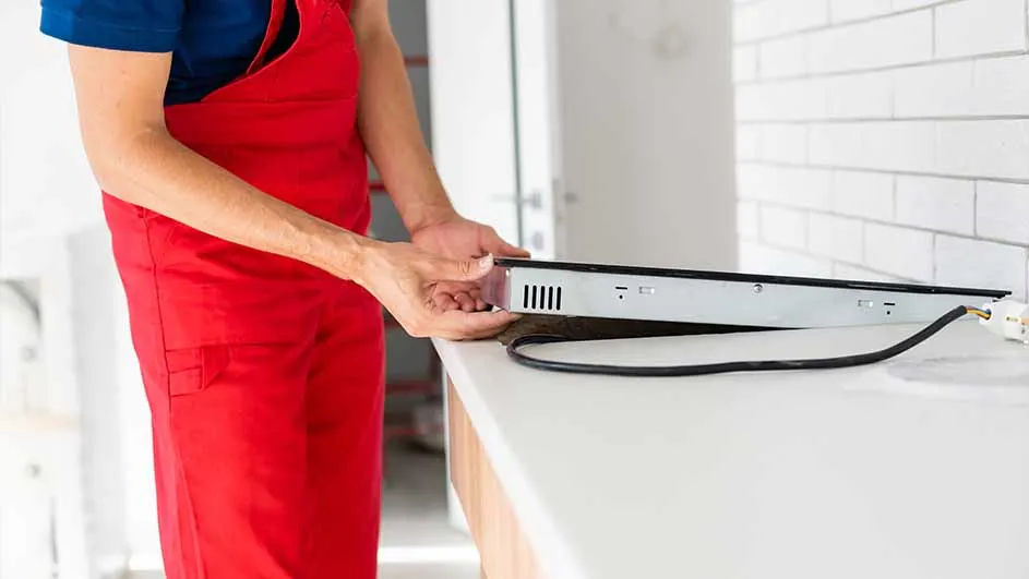 Een persoon in rode overall installeert een zilverkleurige kookplaat op een wit aanrechtblad met een zwarte stroomkabel.