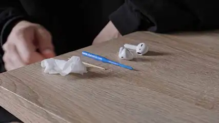 A wooden table holds crumpled tissue, a toothpick, a blue tool, and two white wireless earbuds. A person's hands are visible.