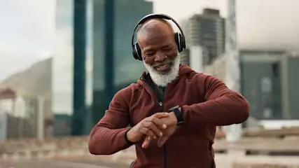 Een man met een baard en een bordeauxrood trainingspak kijkt naar een smartwatch, met een koptelefoon op en gebouwen op de achtergrond.