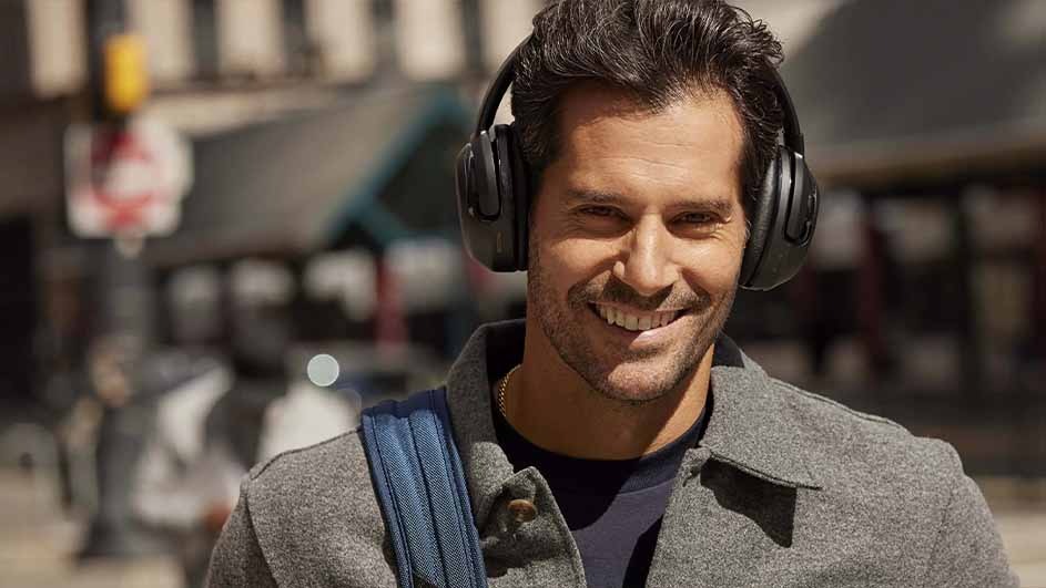 Man smiles wearing headphones, a gray coat, and a blue backpack on a city street.
