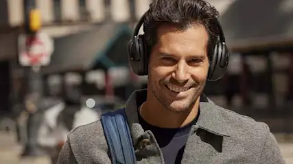 Man smiles wearing headphones, a gray coat, and a blue backpack on a city street.