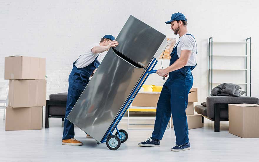 Two movers in blue overalls carefully tilt a tall fridge onto a trolley among cardboard boxes in a sparsely furnished room.