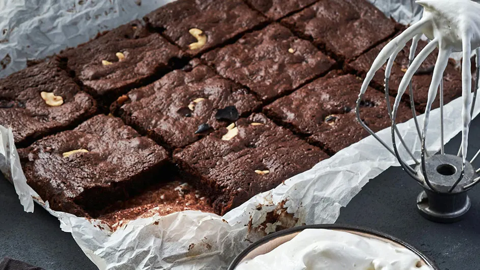 Een bakplaat met vierkante brownies op bakpapier, met een garde en een schaaltje slagroom.