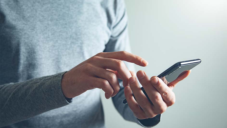 Een persoon met een grijze trui gebruikt een smartphone tegen een witte achtergrond. De persoon houdt de telefoon in de hand en gebruikt de vingers van de andere hand om het scherm aan te raken.