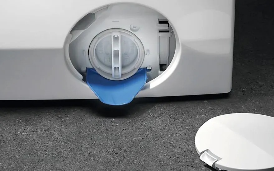 Een open pluizenfilter van een wasmachine met een blauwe handgreep. De ronde witte filterhouder is zichtbaar in de behuizing van de machine. Rechtsonder ligt het losse filterdeksel op een grijze vloer.