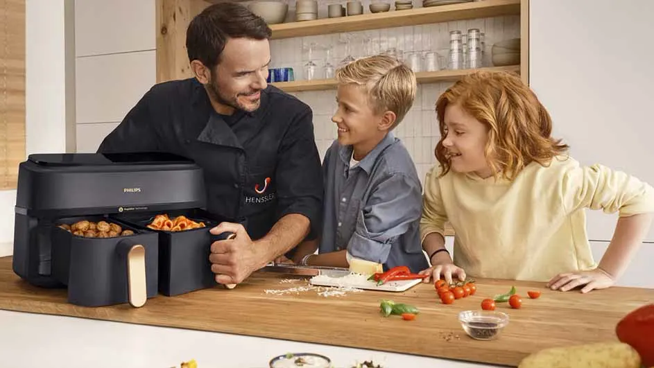 Een man en twee kinderen lachen bij een zwarte Philips airfryer op een houten aanrecht met ingrediënten.