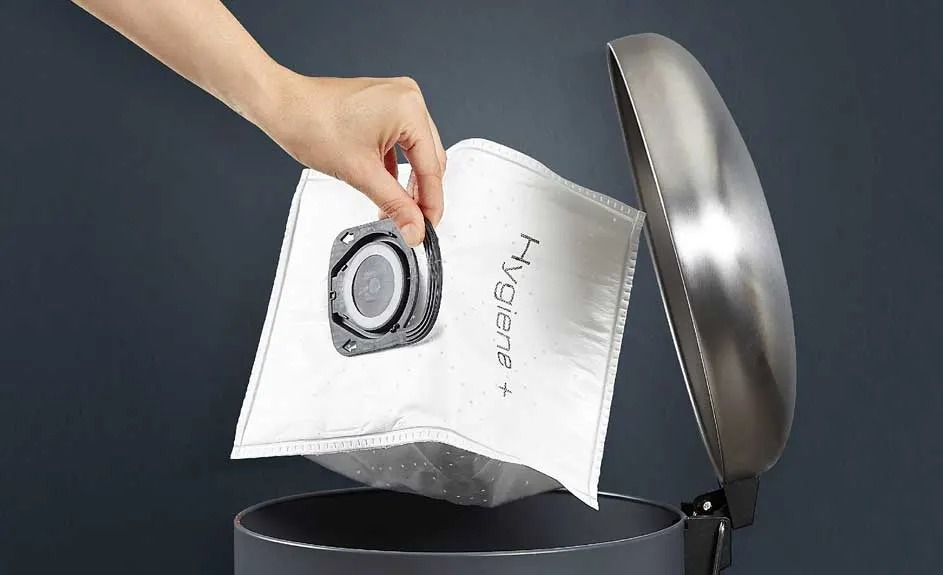 A hand throws a full white vacuum cleaner bag labelled "Hygiene+" into a round, open bin with a metallic lid.
