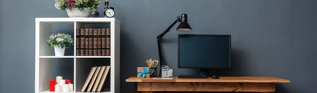 Boekenkast met boeken, bloemen en een wekker naast een bureau met een computer, lamp, potloden en een cadeau tegen een grijze muur.