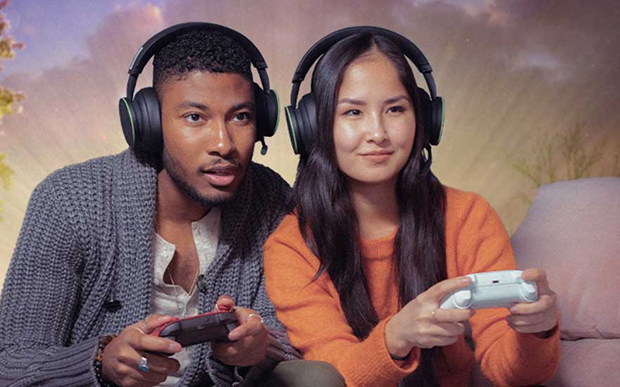 Two young people wearing large headsets game together on a sofa, focused on the screen while each holds a controller in a warm, atmospheric room.
