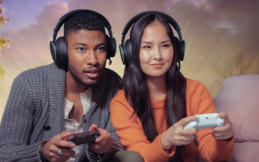 Two young people wearing large headsets game together on a sofa, focused on the screen while each holds a controller in a warm, atmospheric room.