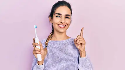 Vrouw lacht, wijst omhoog, houdt witte elektrische tandenborstel vast tegen een paarse achtergrond.