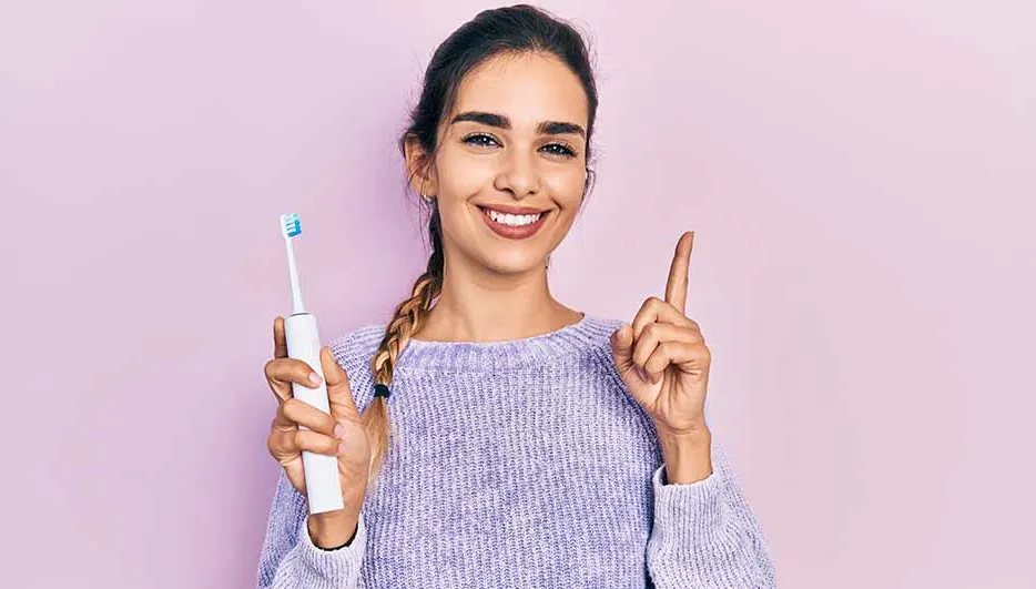Vrouw lacht, wijst omhoog, houdt witte elektrische tandenborstel vast tegen een paarse achtergrond.