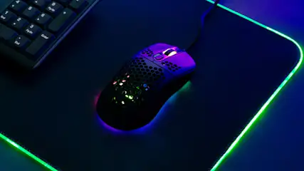 Een zwarte computermuis met paarse accenten ligt op een donkere muismat met groene randverlichting, naast een deel van een toetsenbord.