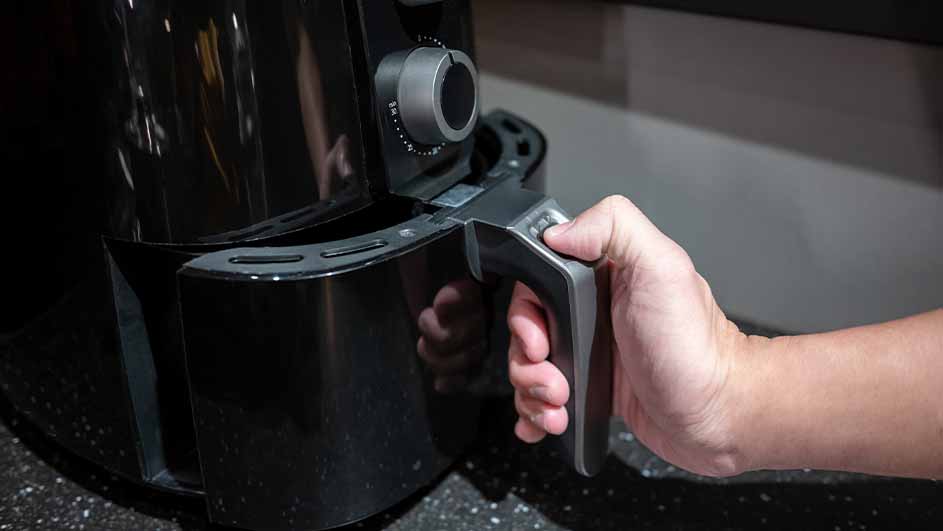 Een hand trekt de lade van een zwarte airfryer open op een aanrecht. De airfryer heeft een zilverkleurige draaiknop.