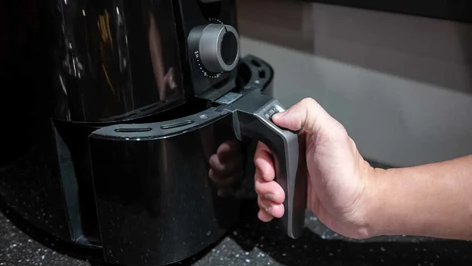 Een hand trekt de lade van een zwarte airfryer open op een aanrecht. De airfryer heeft een zilverkleurige draaiknop.