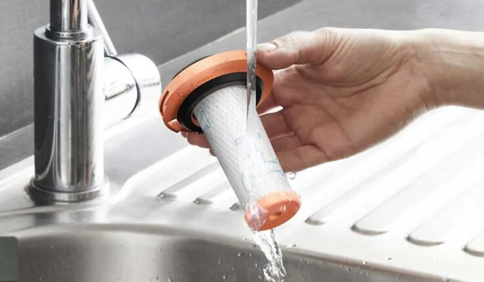 A hand holds a round air filter with an orange edge under running tap water, thoroughly rinsing the filter in the kitchen sink.