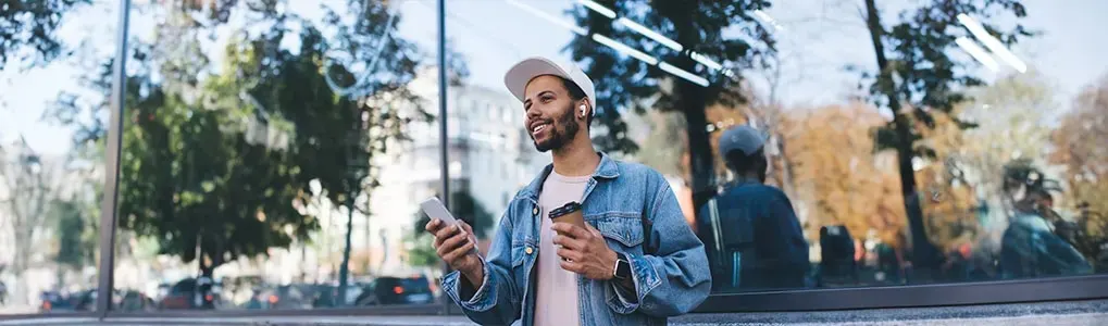 A person in a denim jacket holds a phone and coffee outside a glass building with trees reflected.