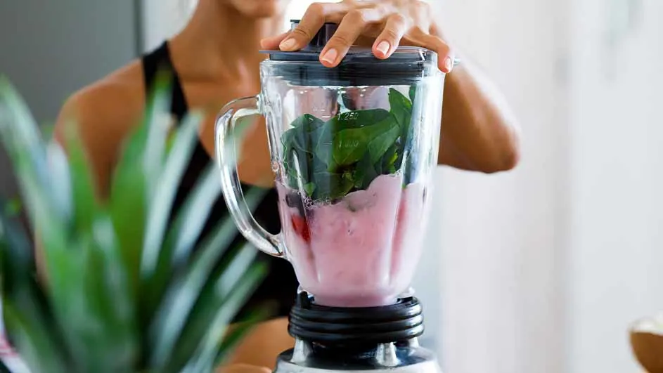 Vrouw maakt smoothie in blender met groene bladeren en roze vloeistof. Plant links.