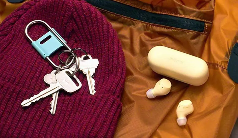 Yellow earbud case with two buds beside a burgundy beanie and keys on orange jacket fabric.