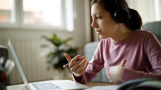 A person wearing headphones and a pink jumper gestures while talking during a video call on a silver laptop, with a plant in the background.