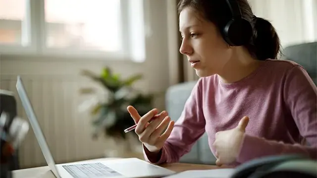 A person wearing headphones and a pink jumper gestures while talking during a video call on a silver laptop, with a plant in the background.