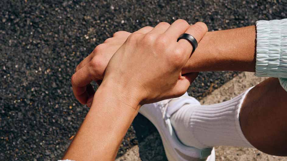 Twee handen, waarvan één met een zwarte ring, rusten op elkaar tegen een achtergrond van asfalt. Witte schoen en sok zichtbaar.