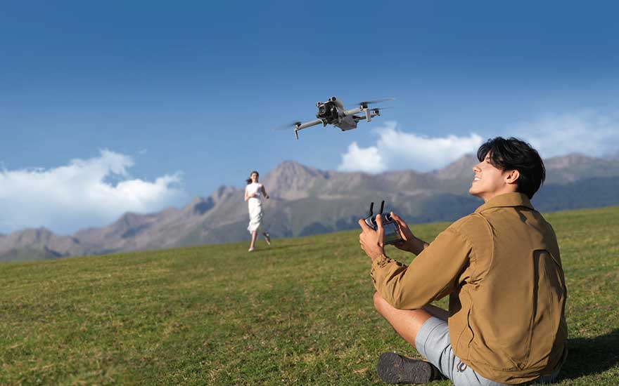 Een man zit op het gras en bestuurt een drone, terwijl op de achtergrond een persoon loopt en bergen zichtbaar zijn. 
