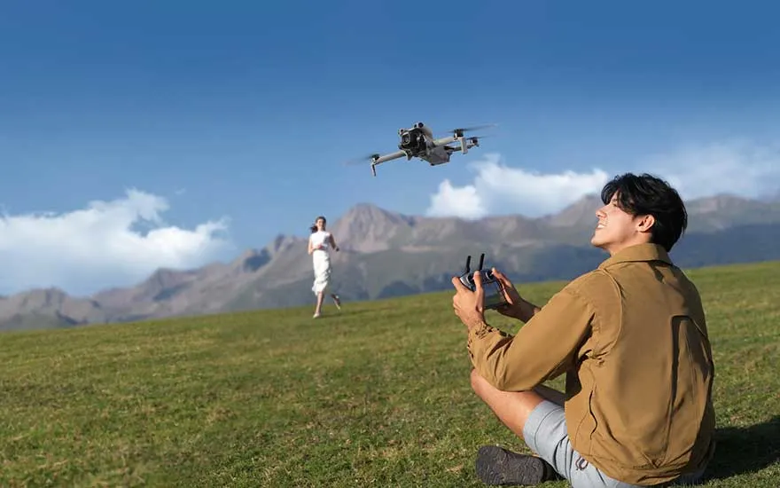 Een man zit op het gras en bestuurt een drone, terwijl op de achtergrond een persoon loopt en bergen zichtbaar zijn.