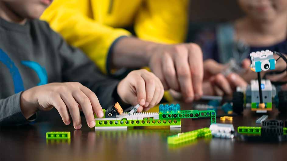 Kinderen spelen met LEGO-stenen op een tafel. Ze bouwen een robot met groene, witte en blauwe blokken.