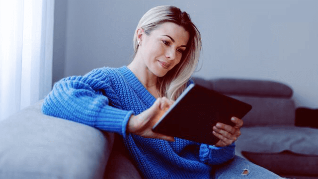 Vrouw in blauwe trui zit op een grijze bank en gebruikt een zwarte tablet. Achtergrond: een raam met witte gordijnen.
