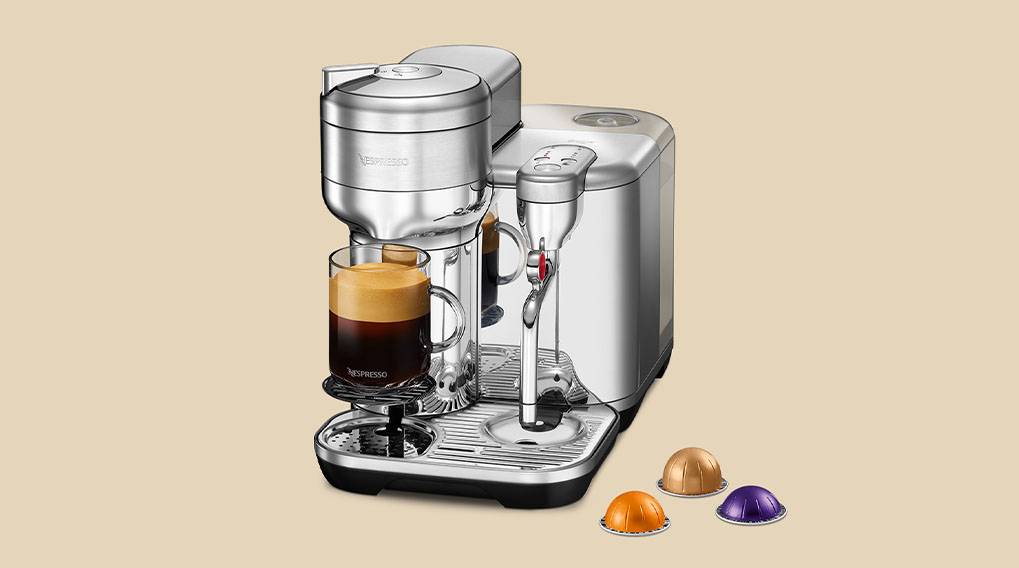 Een zilverkleurige Nespresso-machine met een glas koffie, geplaatst op een beige achtergrond met koffiecapsules ervoor.