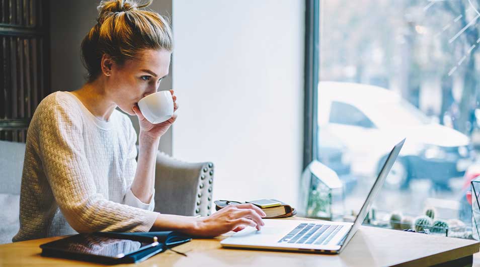 Vrouw drinkt koffie en werkt geconcentreerd op haar laptop bij het raam in een gezellige ruimte.