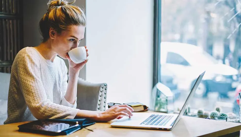 Vrouw drinkt koffie en werkt geconcentreerd op haar laptop bij het raam in een gezellige ruimte.