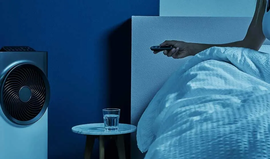 Een persoon ligt in bed en gebruikt een afstandsbediening voor een Dyson ventilator, naast een glas water op een tafeltje.