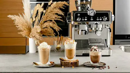 Drie glazen koffie met slagroom, naast een zilverkleurige espressomachine en een vaas met pluimen.