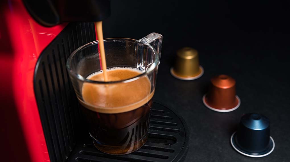 Koffie wordt in een glazen kopje gegoten door een rood koffiezetapparaat, met capsules goud, oranje en blauw op een zwarte ondergrond.