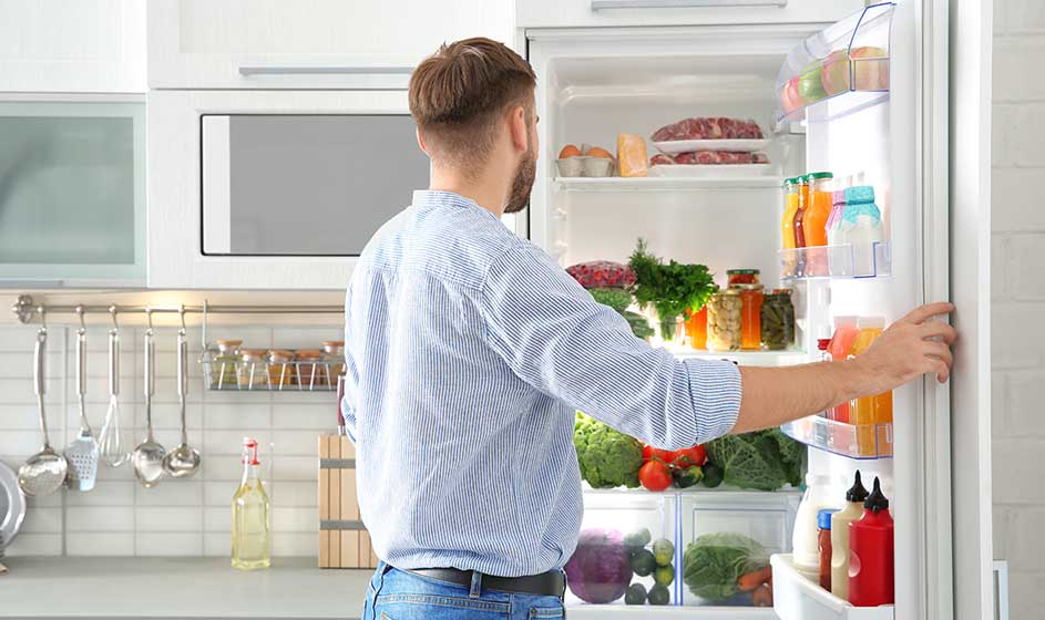 A man in a bright kitchen looks inside an open fridge full of vegetables, fruit, jars and bottles while holding the door.