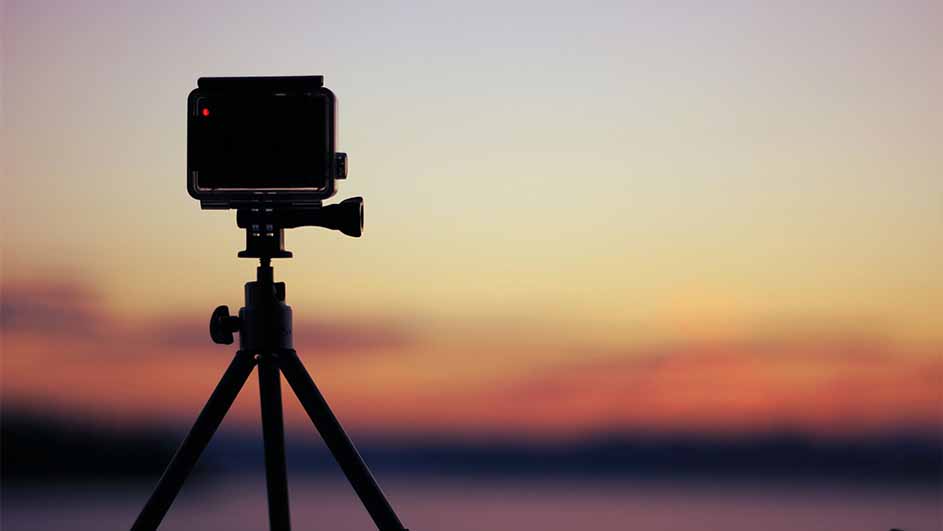 Actiecamera op statief tegen zonsondergang met oranje, gele en blauwe luchtkleuren.