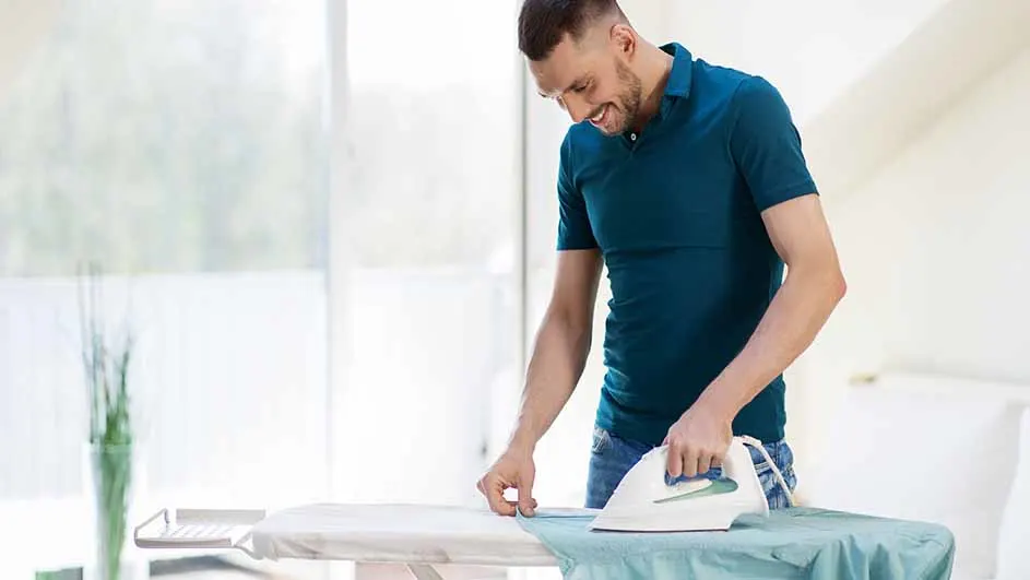 Een man strijkt een lichtblauw kledingstuk op een strijkplank. Hij draagt een blauw poloshirt en jeans. Achtergrond: een raam en planten.