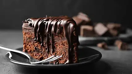 Een stuk chocoladetaart met glazuur op een zwart bord met een zilveren vork. Chocoladeblokjes op de achtergrond.