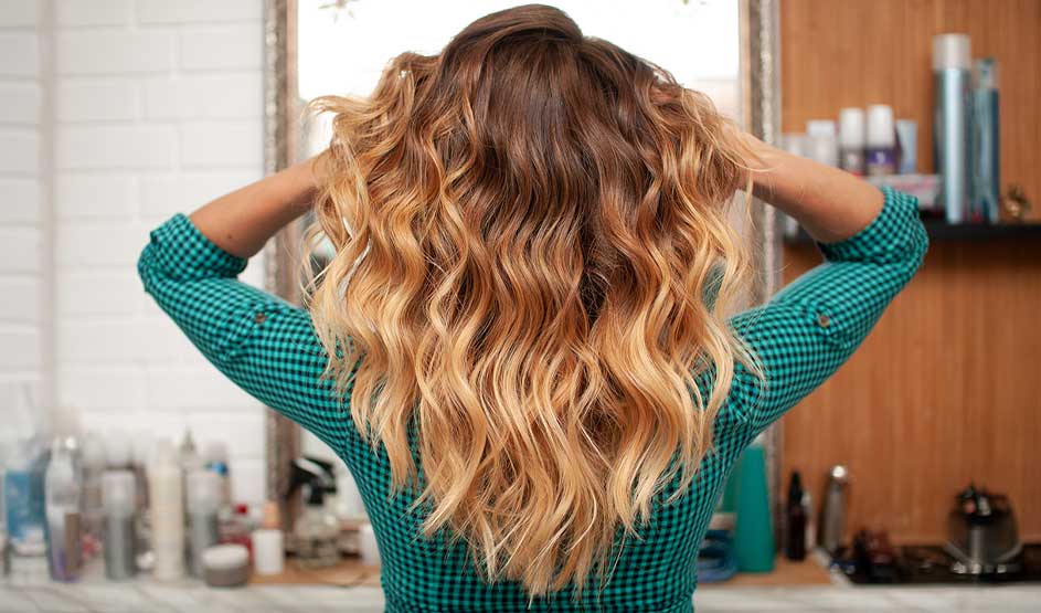 A person seen from behind shows long, wavy ombré hair in a salon, hands in their hair, with styling products on shelves.
