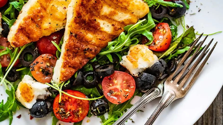 Een bord met gegrilde kaas, rucola, rode tomaten en zwarte olijven. Een vork ligt aan de rand van het bord.