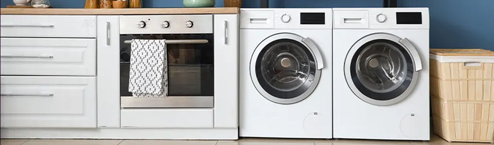 A kitchen with white cabinets, a stainless steel oven with a towel, two white washing machines, and a wicker basket.