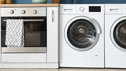 A kitchen with white cabinets, a stainless steel oven with a towel, two white washing machines, and a wicker basket.