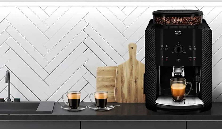 Op een modern aanrecht staat een zwarte Krups koffiemachine met koffiebonen, drie glazen koffie en twee houten snijplanken op de achtergrond.