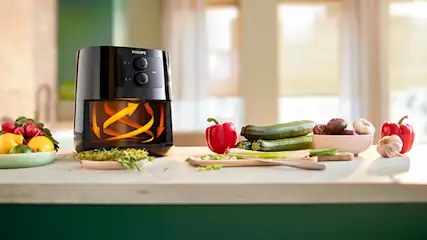 Een zwarte Philips airfryer staat op een aanrecht met groenten en fruit. Er staan rode paprika's, courgettes, uien en knoflook.