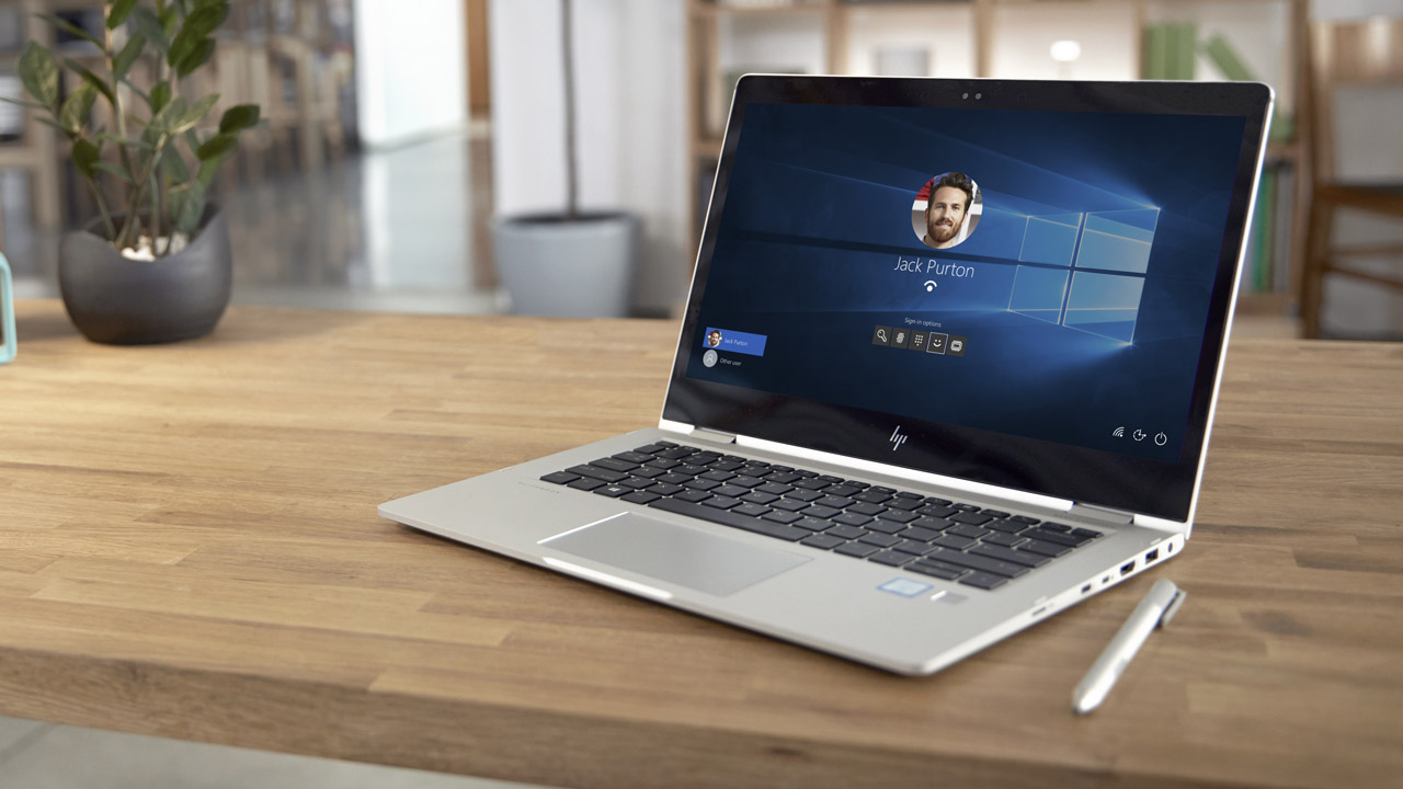 Een zilveren laptop staat open op een houten bureau met een Windows-aanmeldscherm en een zilveren stylus.